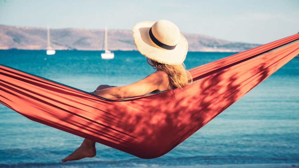 Άτομο χαλαρώνει σε αιώρα δίπλα στη θάλασσα, φορώντας καπέλο